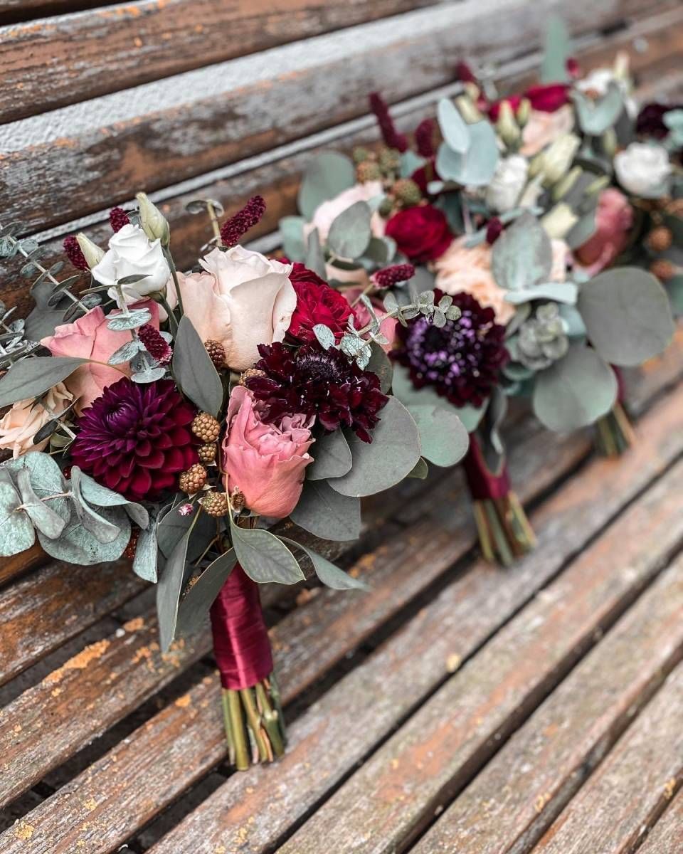 Drei Brautsträusse aus tief burgunderroten Dahlien, rosa Rosen und Eukalyptusgrün, mit Bändern auf einer Holzbank zusammengebunden.