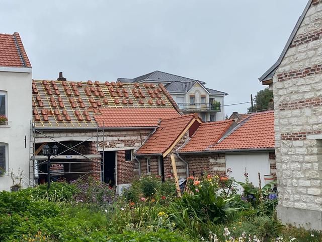 Bâtiments aux toits de tuiles rouges, certains en construction. Feuillage vert au premier plan. Ciel couvert.