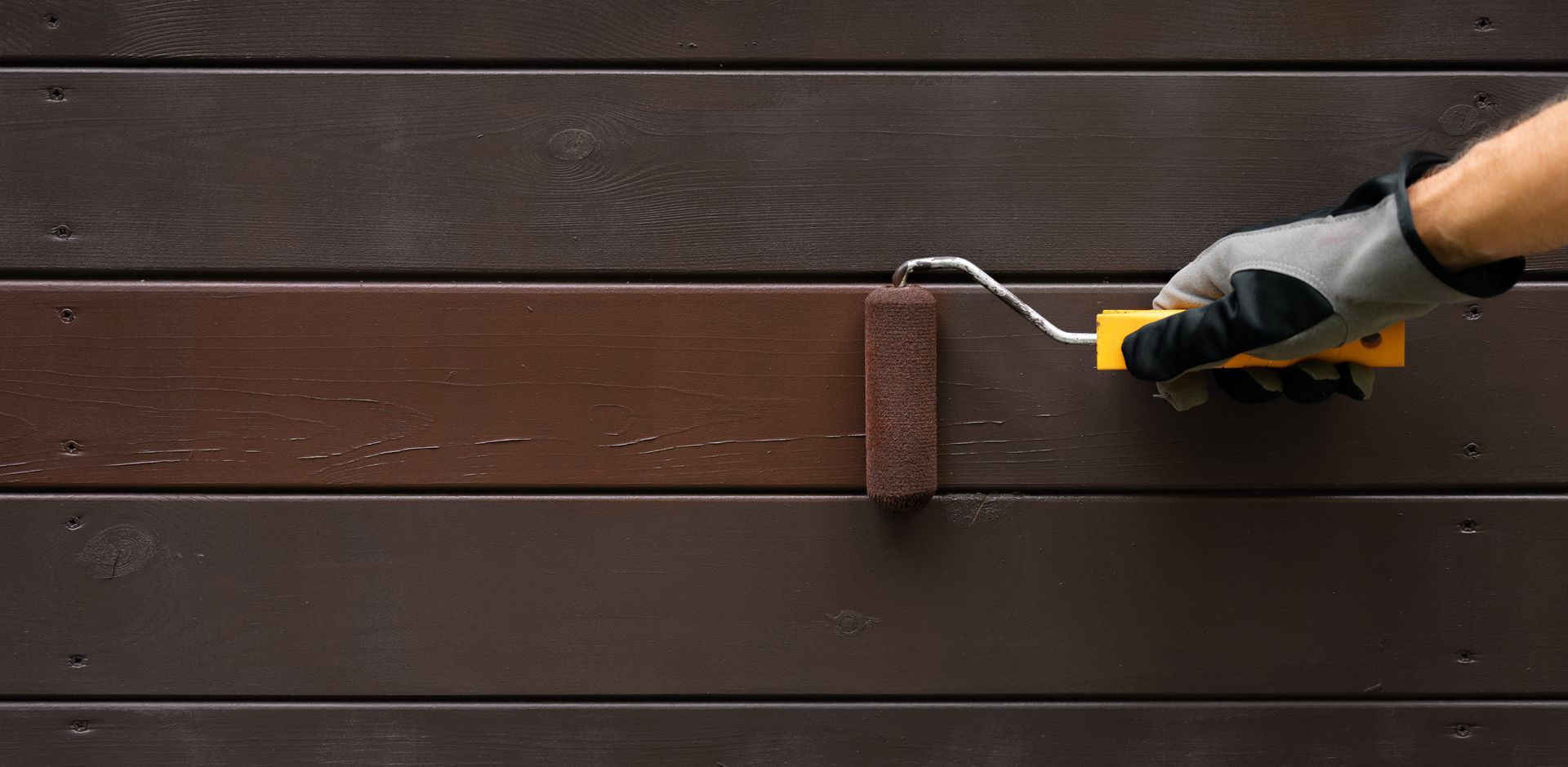 Une personne peint une clôture en bois avec un rouleau à peinture, appliquant de la peinture marron.