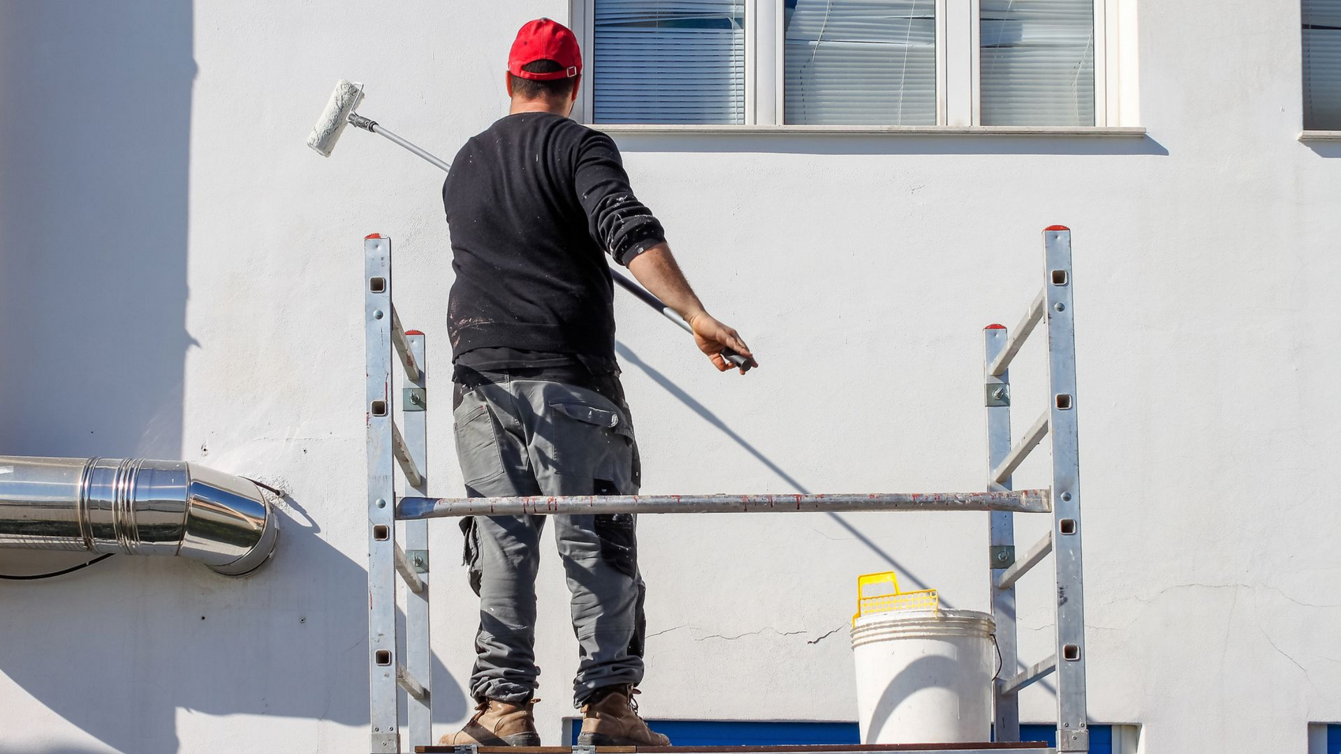 Un homme peint un mur extérieur debout sur un échafaudage, à l'aide d'un rouleau.