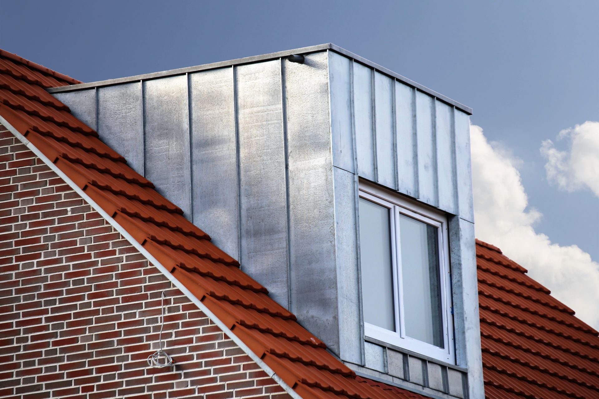 Lucarne avec revêtement en métal galvanisé sur toit en tuiles rouges contre un ciel bleu.