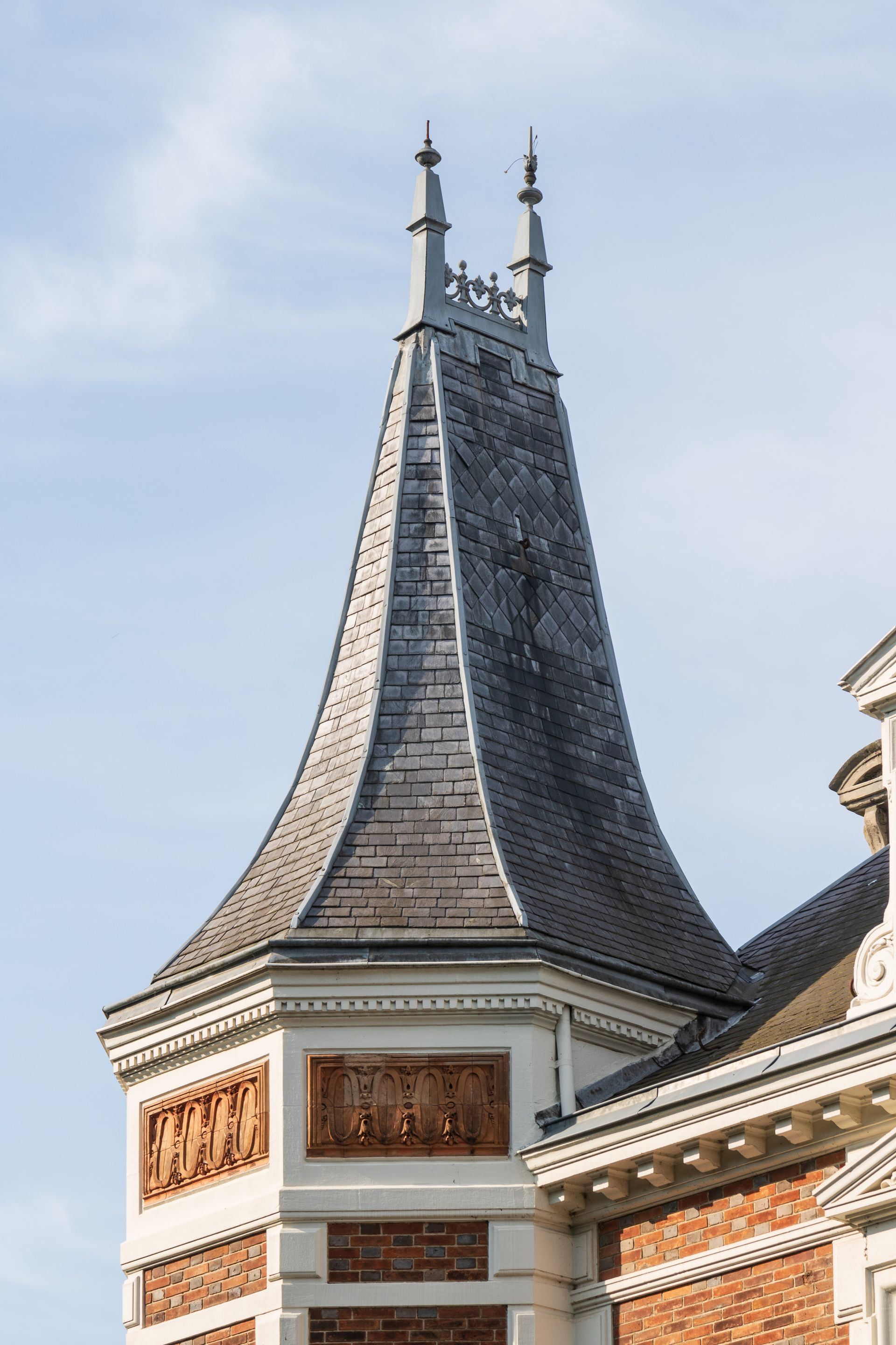 Tour avec un toit pointu en bois et une flèche décorative, partie d'un bâtiment en brique et en pierre, contre un ciel bleu.