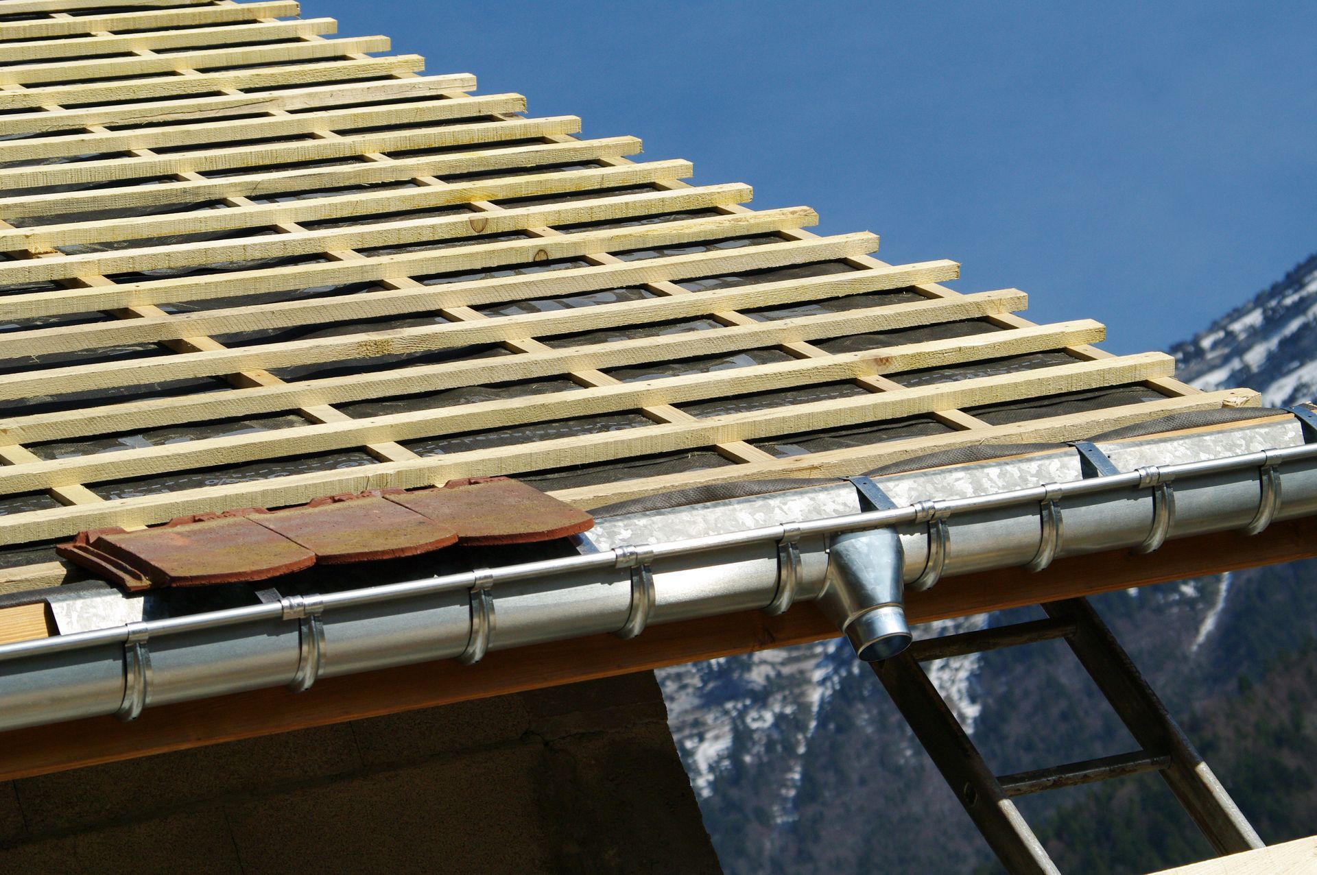 Toiture en construction, avec charpente en bois, isolation et gouttière, sur fond de montagne.