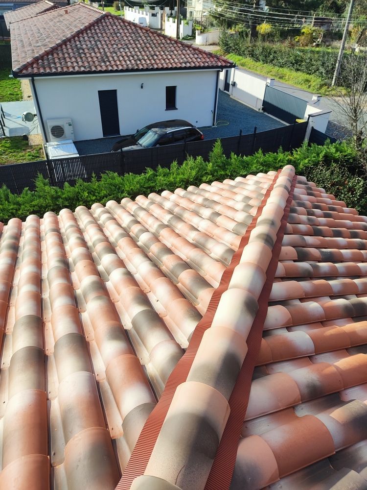 Gros plan sur un toit de tuiles dans les tons bruns et rouges, avec une maison en arrière-plan.