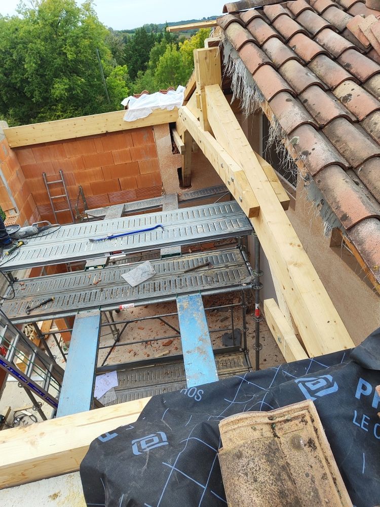 Chantier : poutres en bois, mur de briques, toit de tuiles, échafaudages, isolation bleue, papier goudronné et arbres.