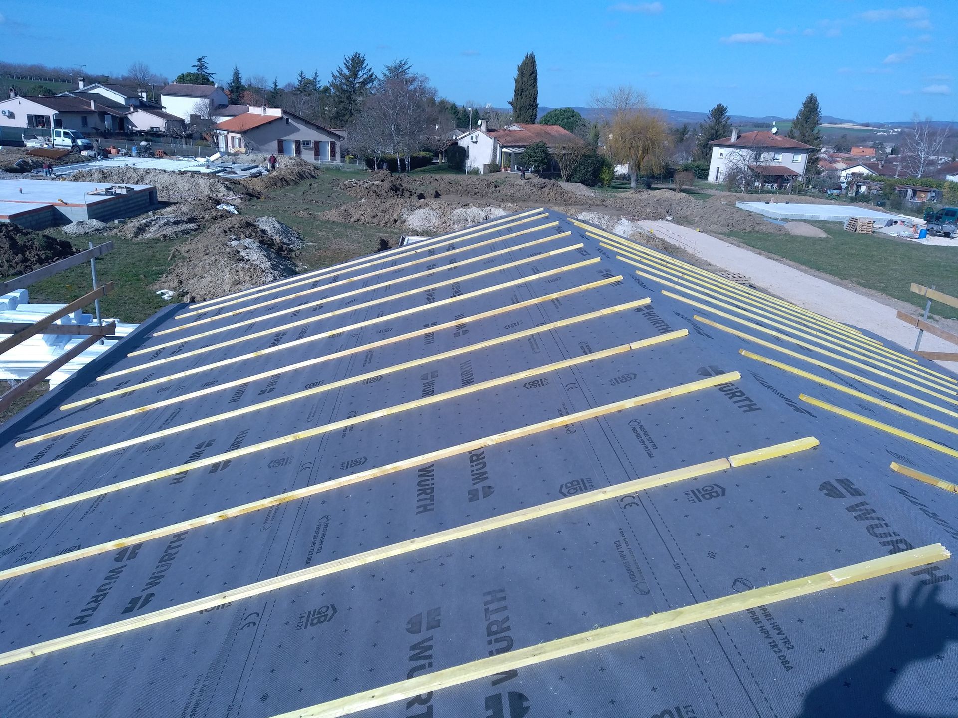 Toiture partiellement construite avec sous-couche noire et liteaux en bois, zone résidentielle en arrière-plan.