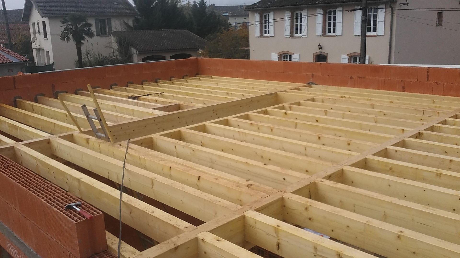 Des poutres en bois forment la structure du plancher, entourées de murs en briques, avec des bâtiments en arrière-plan.