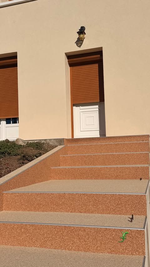 Des marches en pierre mènent à un bâtiment beige avec une porte blanche et des volets marron.