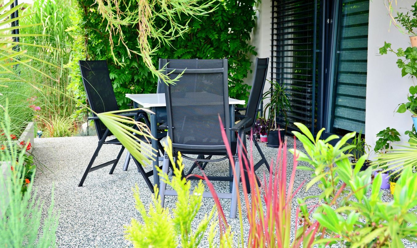 Table et chaises de patio noires sur une terrasse en gravier, à côté d'une végétation luxuriante et de la façade d'une maison.