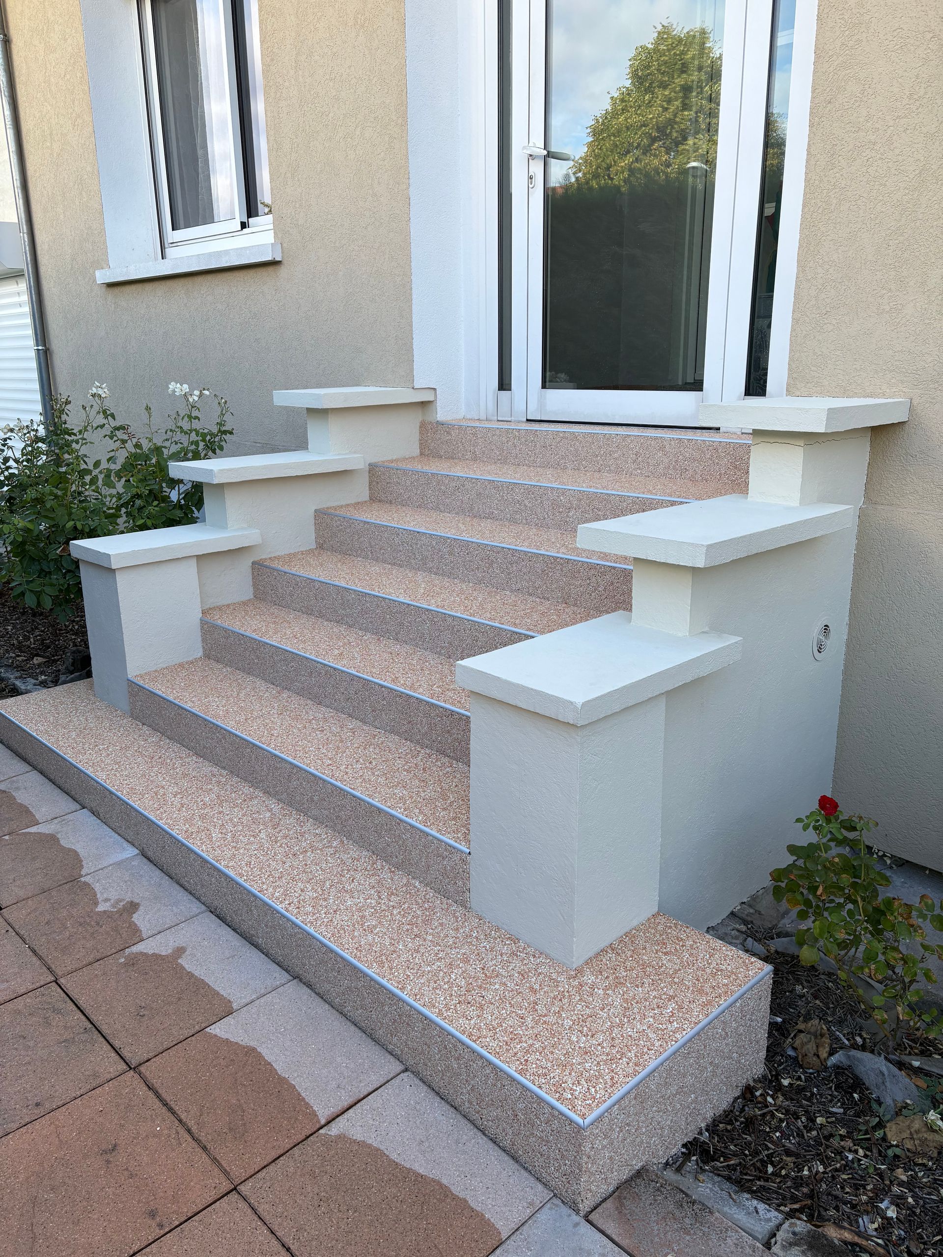 Des marches en pierre mènent à un bâtiment beige avec une porte blanche et des volets marron.
