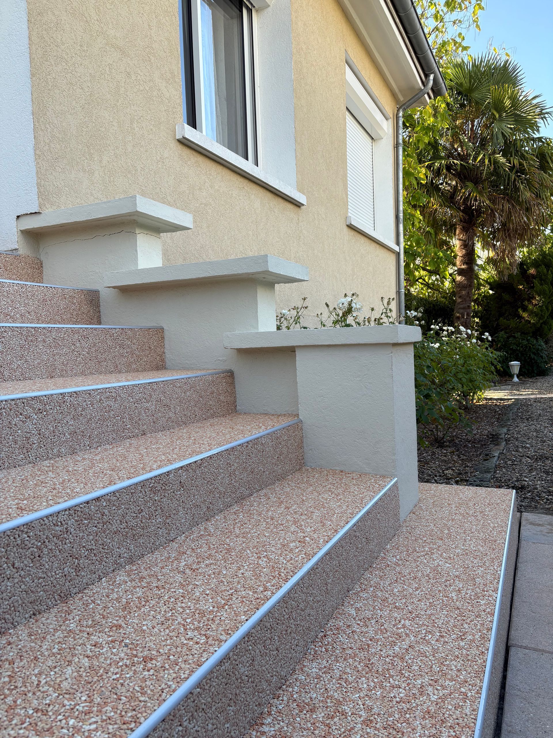 Escaliers extérieurs en pierre jouxtant une maison couleur crème, avec un petit palier et un chemin de jardin ensoleillé.