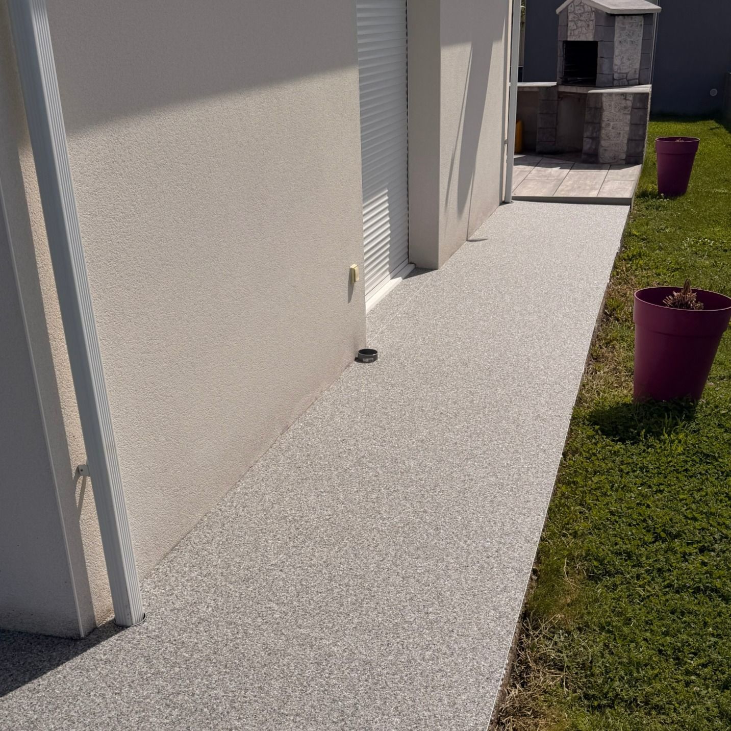 Un étroit chemin de béton longe une maison, bordé de pelouse et de jardinières violettes baignées de soleil.