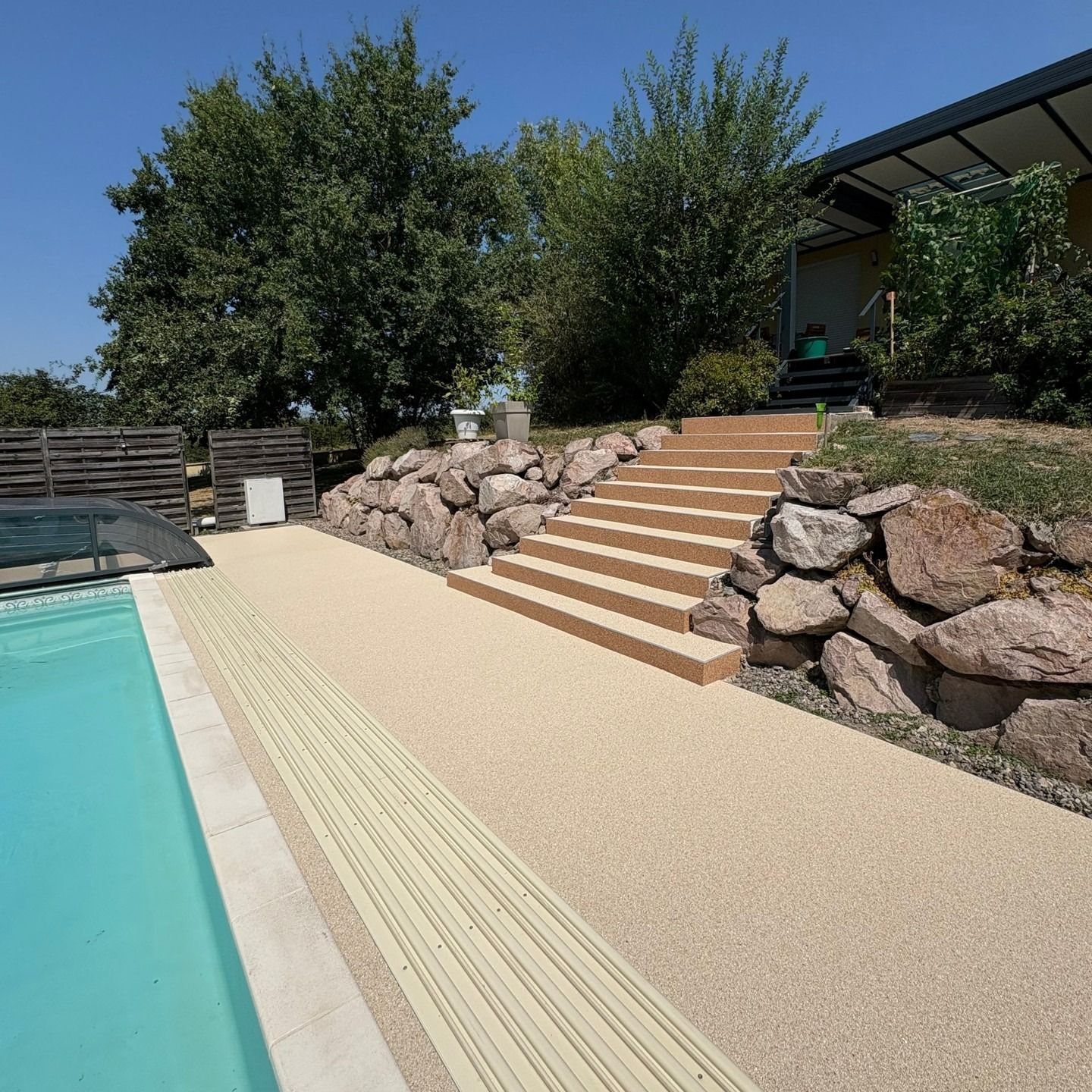 Piscine dans le jardin avec terrasse en galets beiges, marches en pierre et pente aménagée à côté d'une maison