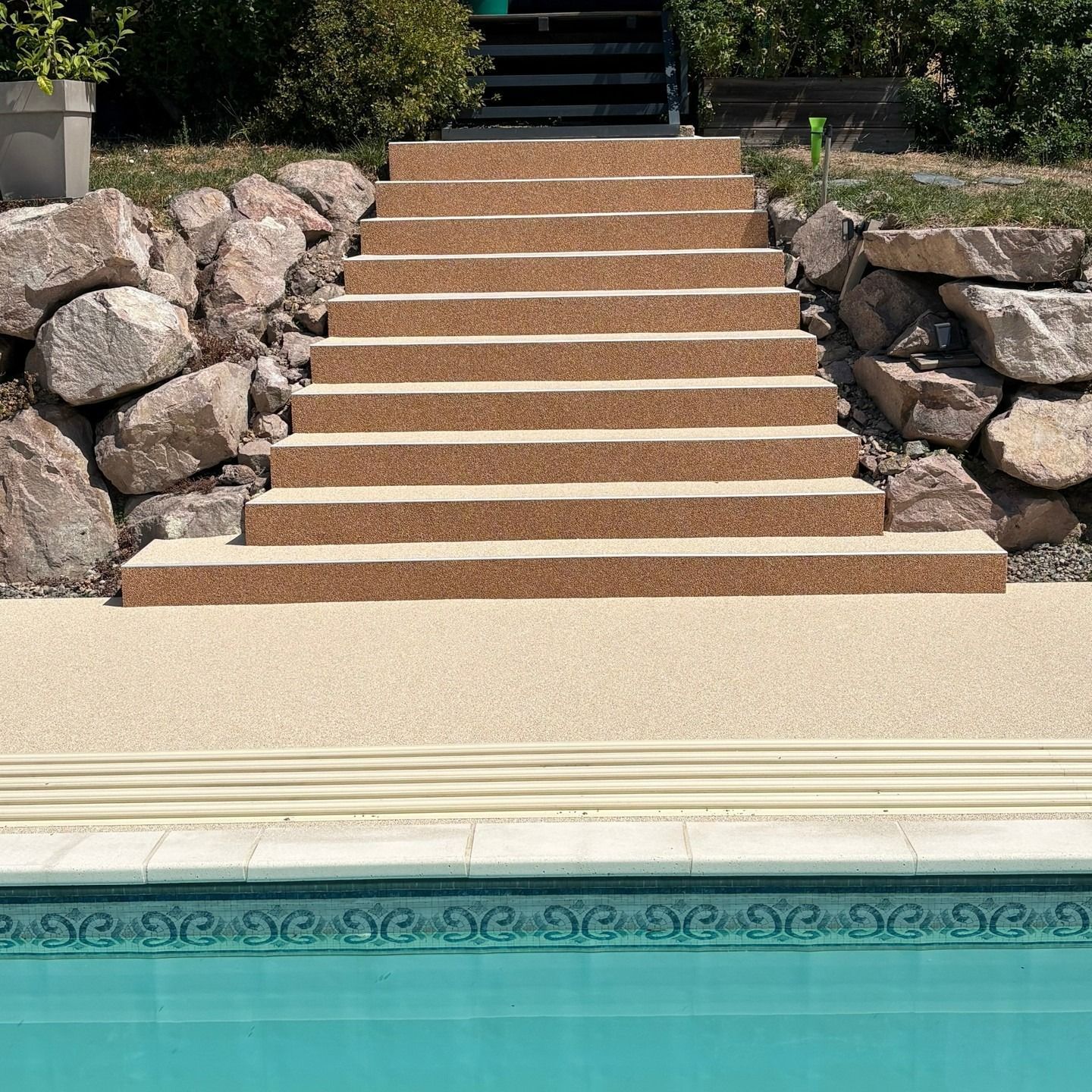 Des marches en pierre entre les rochers mènent à une piscine turquoise ornée de carreaux décoratifs.