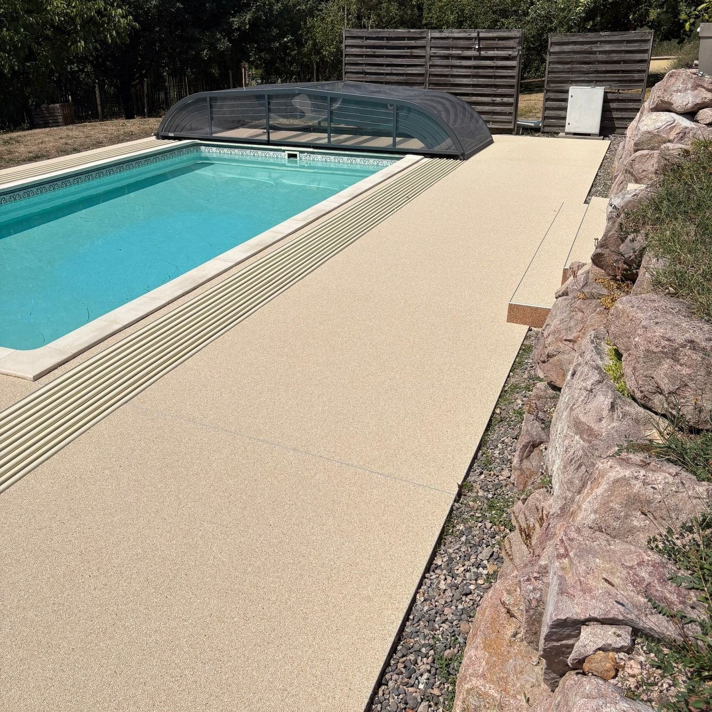 Piscine de jardin avec terrasse texturée beige, couverture de sécurité et aménagement paysager rocailleux.
