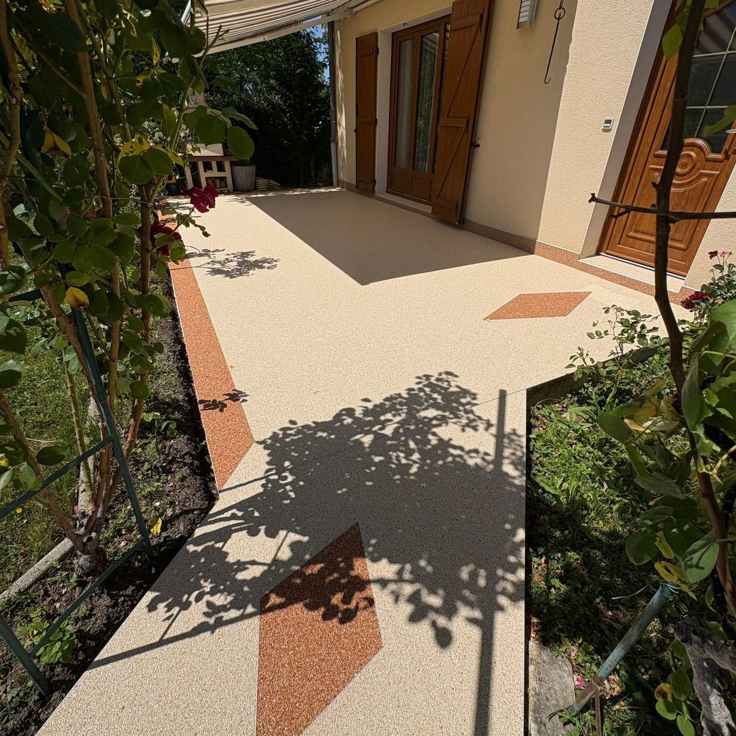 Allée carrelée et ensoleillée longeant une maison, bordée de plantes de jardin et de portes en bois.