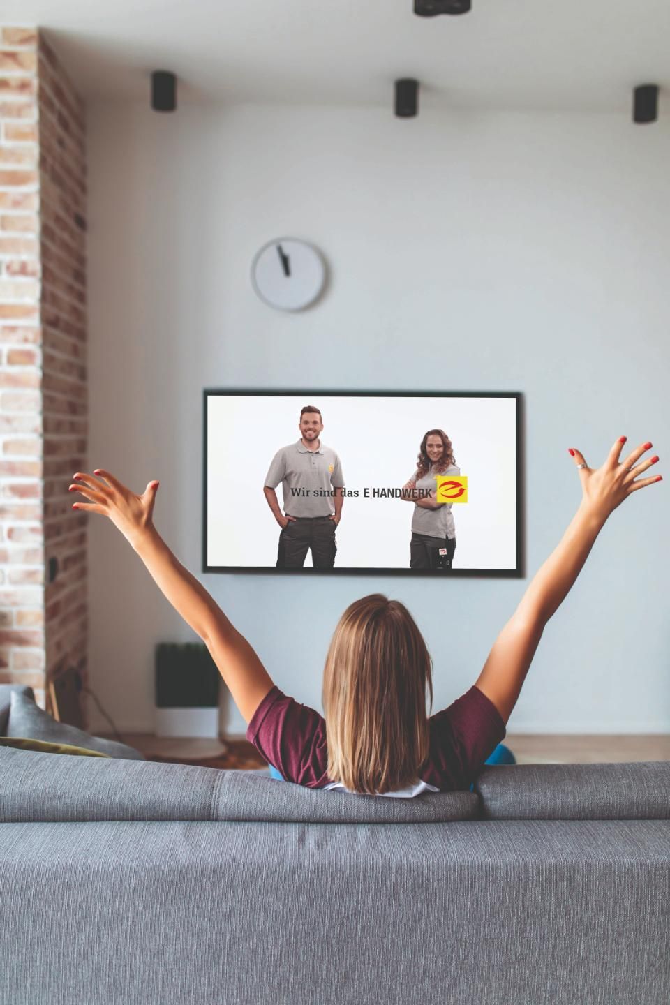 Rückenansicht von auf grauer Couch sitzender braunhaarigen Frau, die beide Arme in die Luft streckt, und auf einen Fernseher mit dem Logo E-Handwerk blickt