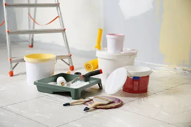 Suministros de pintura, incluidos baldes de pintura, rodillo y escalera, en una habitación con pared pintada.