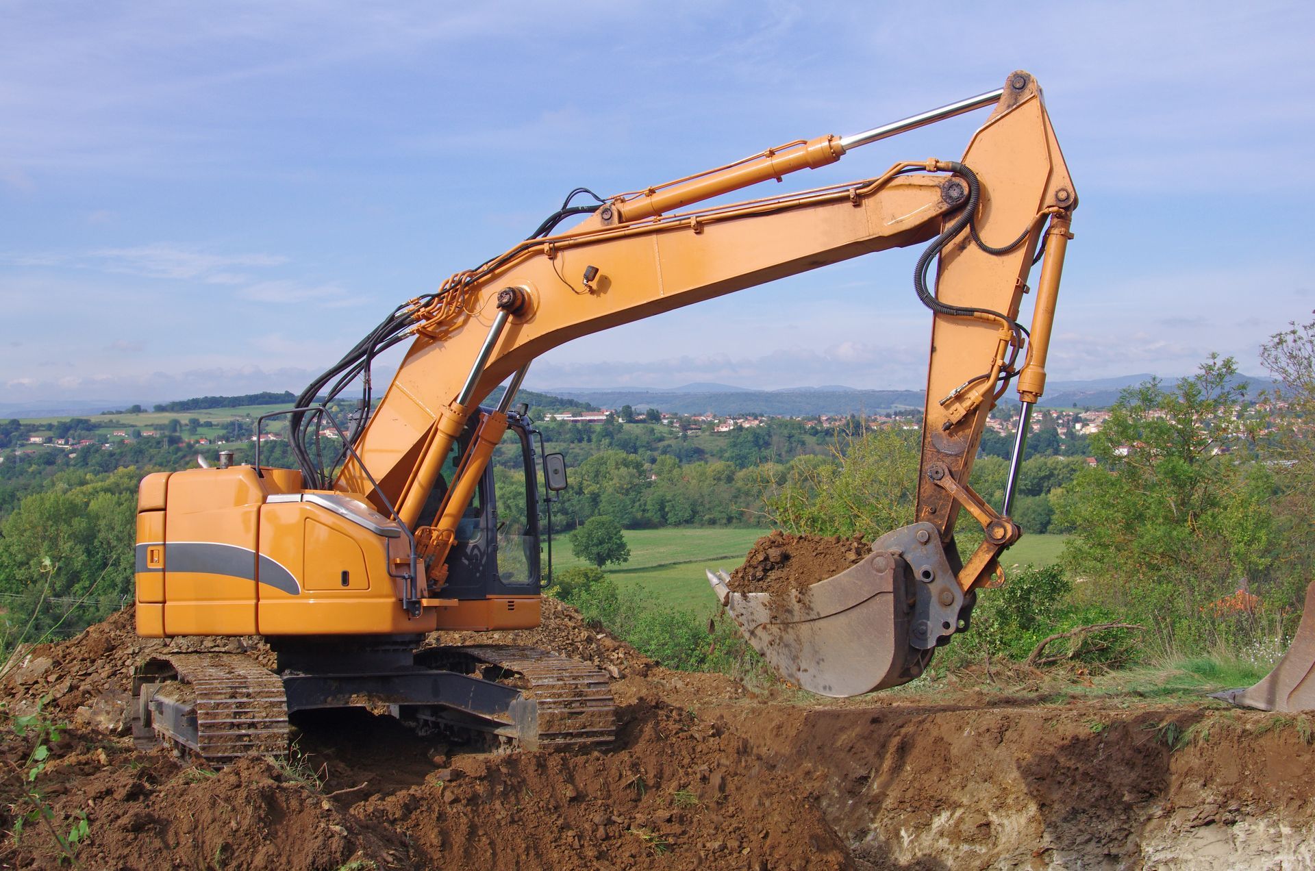 Une pelleteuse orange creuse la terre sur un chantier de construction, avec un paysage verdoyant en arrière-plan.
