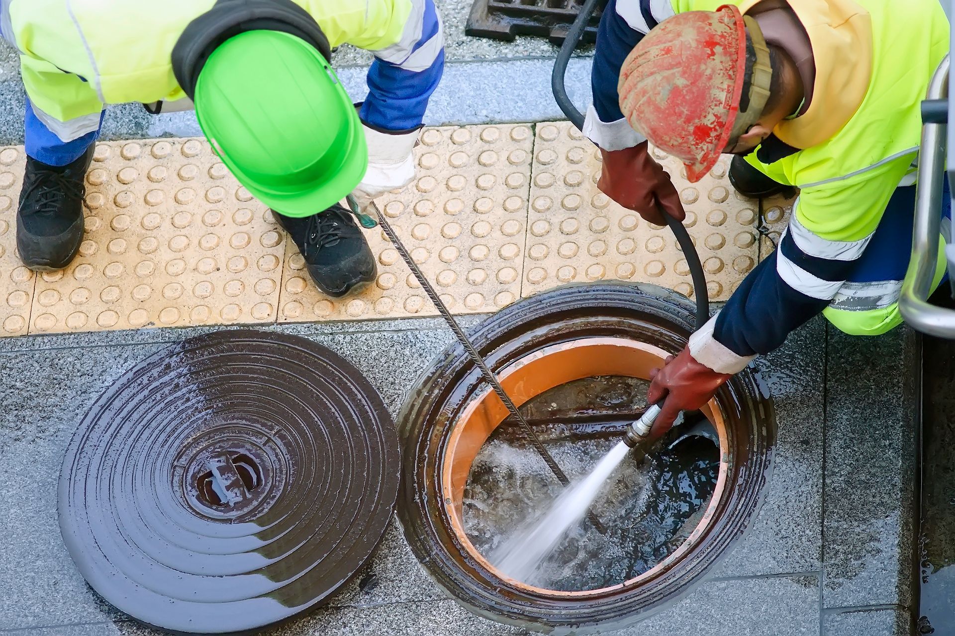 Deux ouvriers nettoient un regard d'égout avec de l'eau, portant des gilets de sécurité et des casques, sur un trottoir.