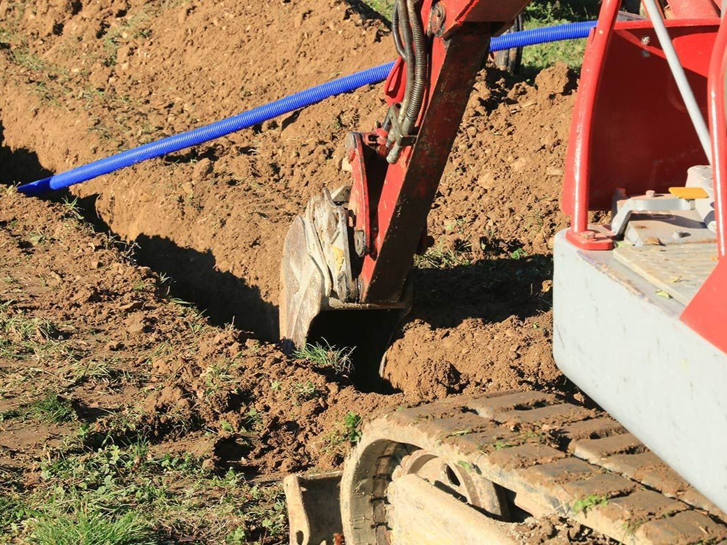 Une excavatrice creuse une tranchée, un tuyau bleu est visible.