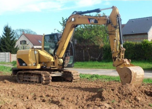 Excavateur Caterpillar jaune creusant dans une zone de terre, avec des maisons en arrière-plan.