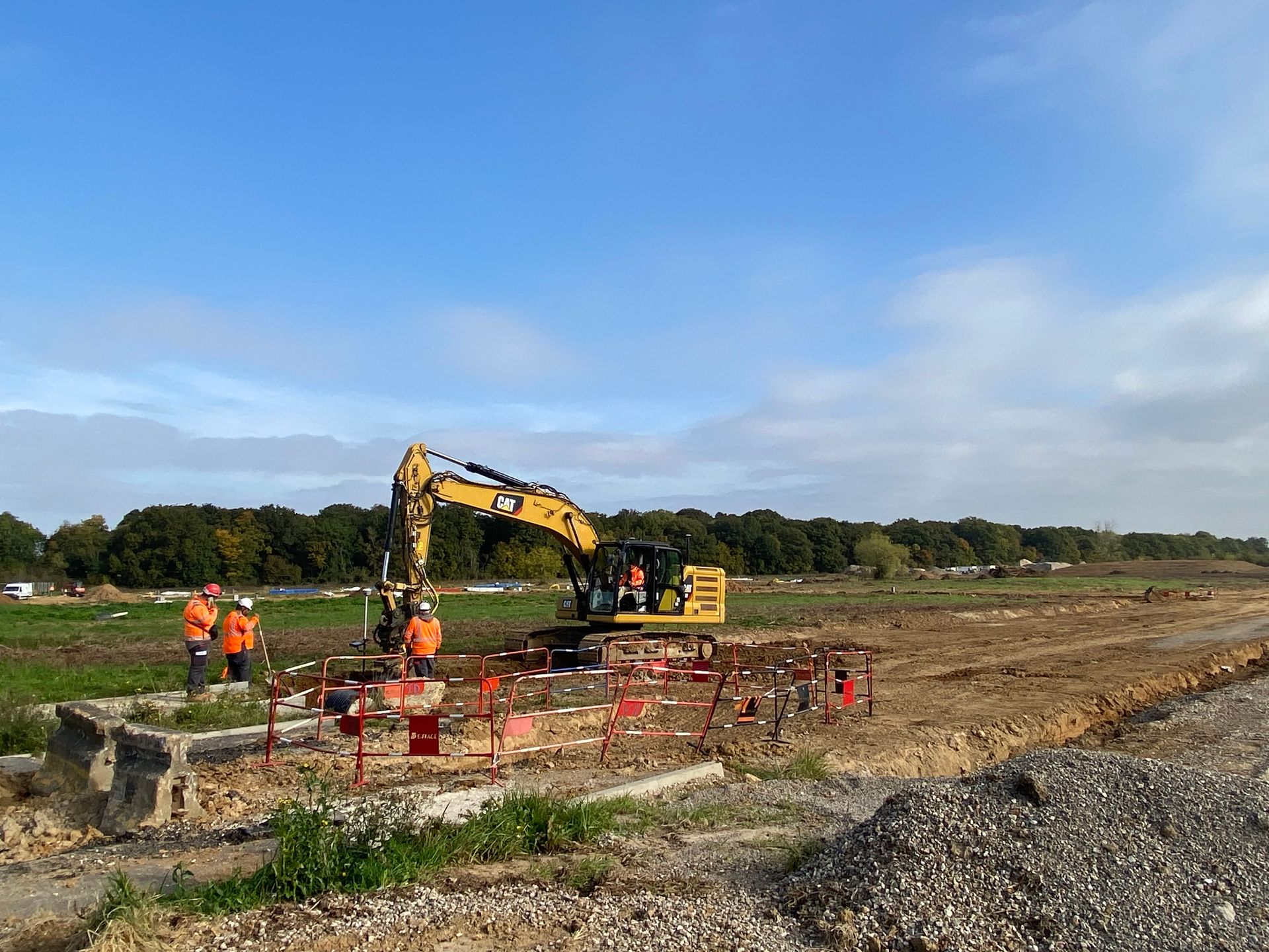 Chantier de construction avec une excavatrice, des ouvriers en gilet orange et un ciel bleu dégagé.