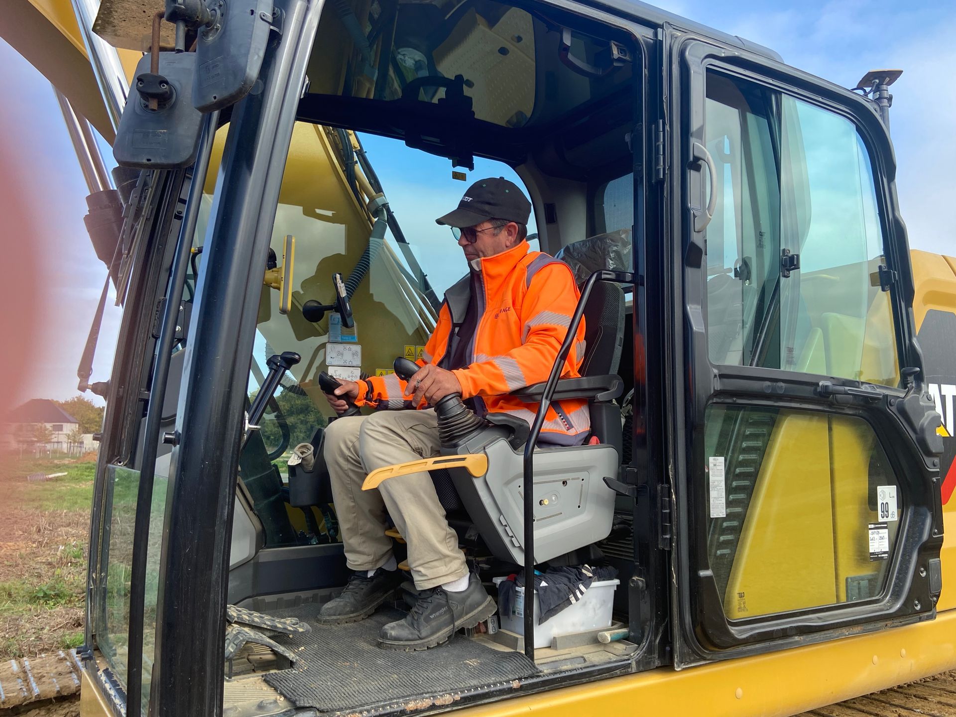 Une personne manœuvrant une pelle mécanique de chantier, portant un gilet de sécurité et une casquette orange, à l'extérieur.