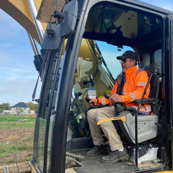 Un homme portant un gilet de sécurité orange manœuvre une excavatrice dans un champ.