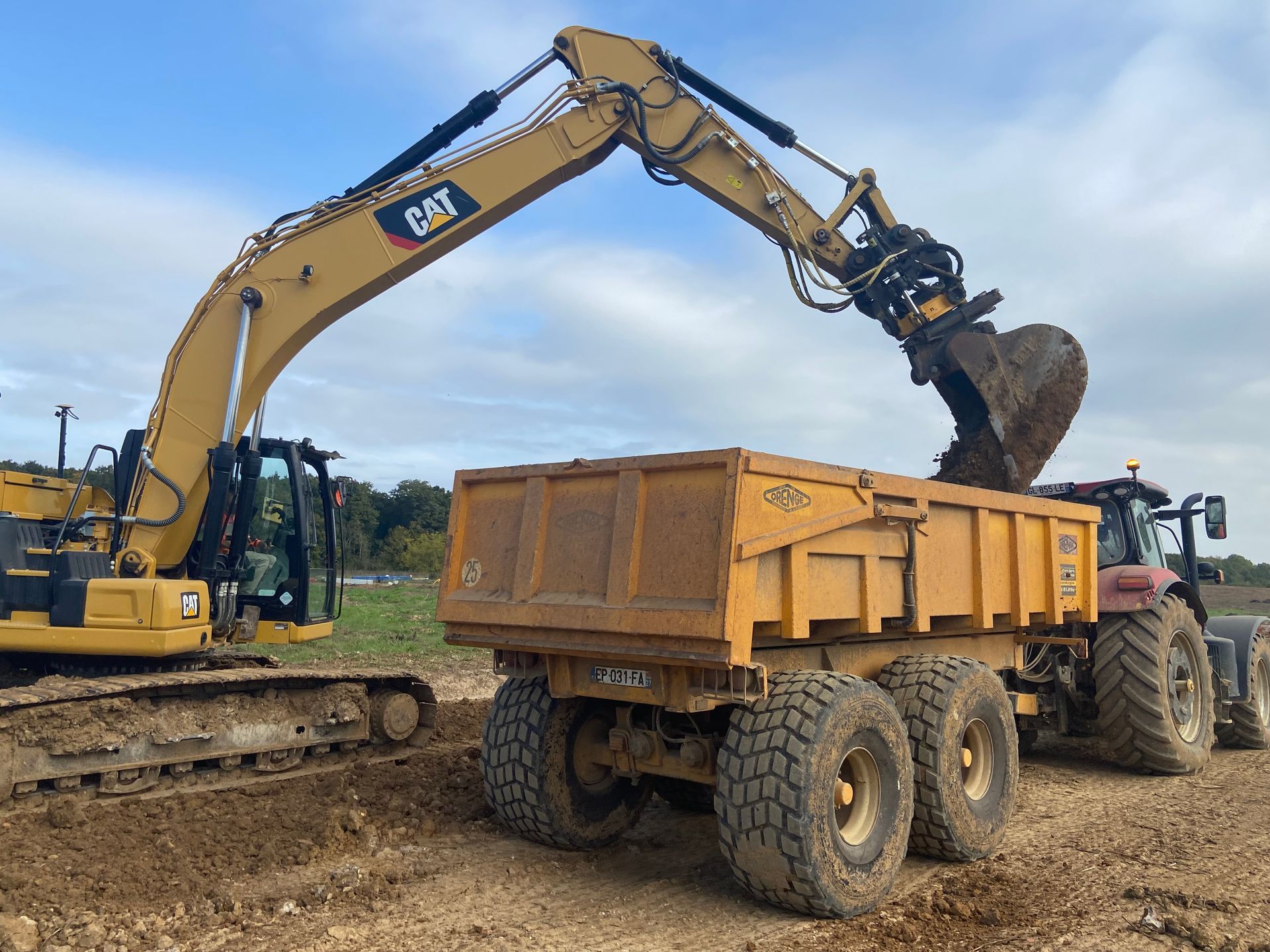 Une pelle mécanique jaune charge de la terre dans une benne basculante jaune tirée par un tracteur, dans un champ.