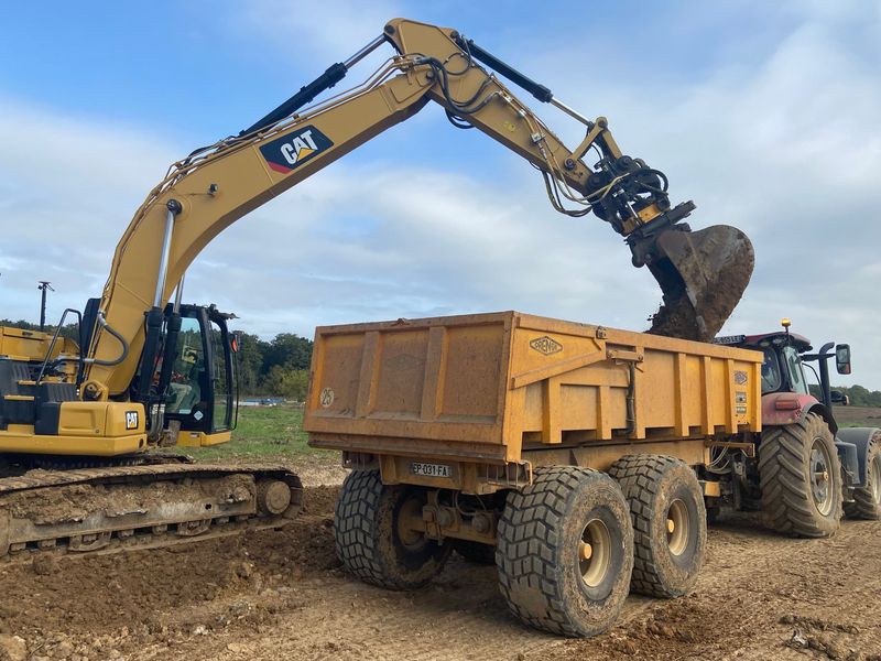 Une pelle mécanique jaune charge de la terre dans une benne basculante jaune tirée par un tracteur, dans un champ.