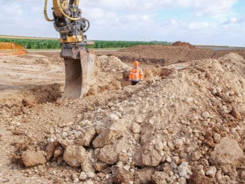 Un ouvrier du bâtiment en gilet orange près d'une excavatrice et d'un tas de terre dans un champ.