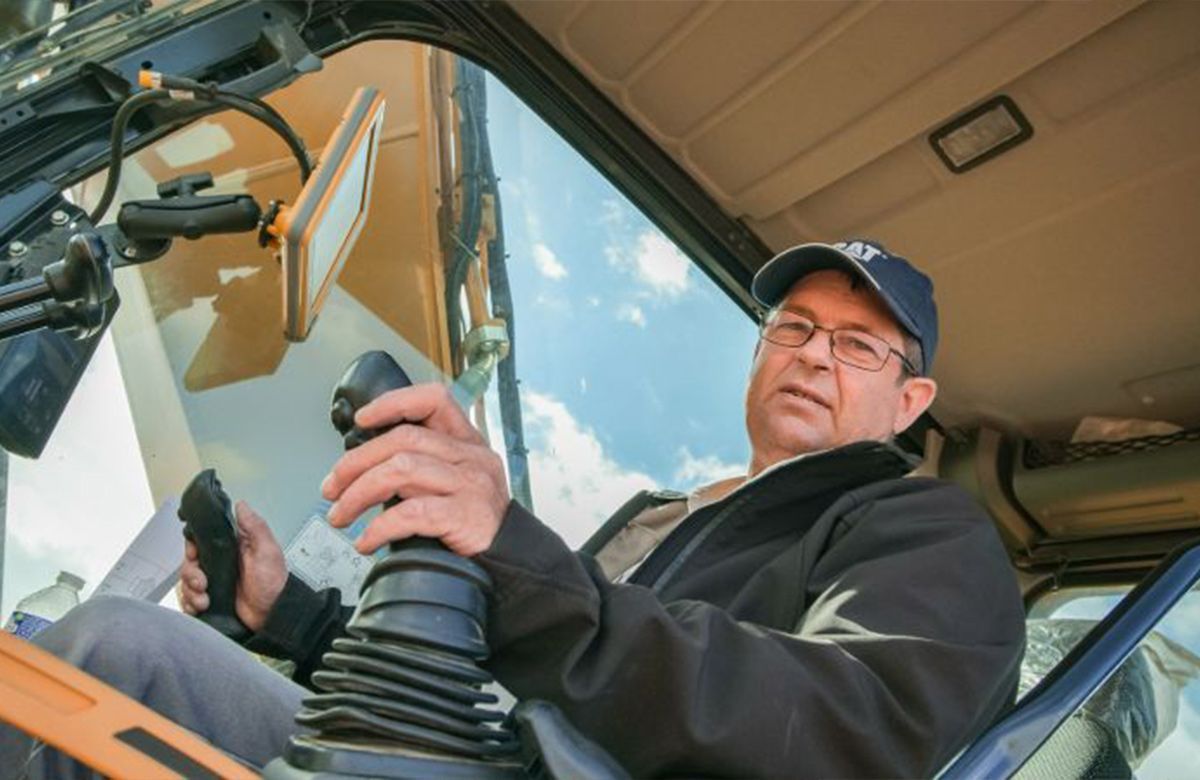 Un homme manœuvre une machine, tenant le manche à balai. Le ciel bleu est visible par la fenêtre de la cabine.