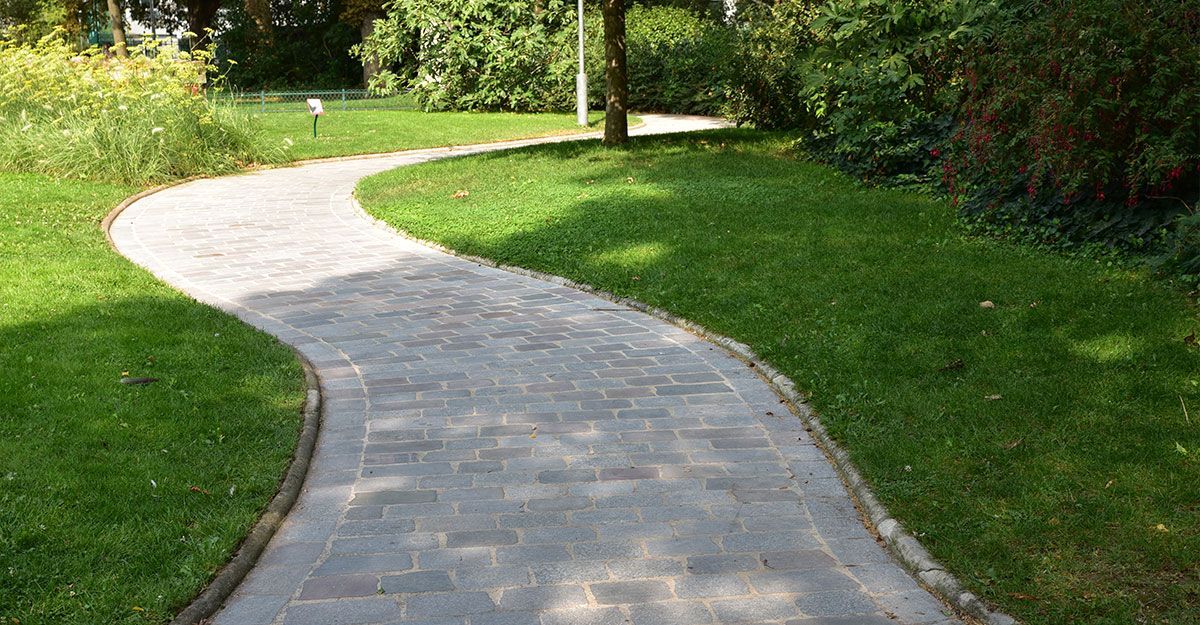 Un chemin de pierre serpente à travers l'herbe verte d'un parc, avec des arbres et des buissons en arrière-plan.
