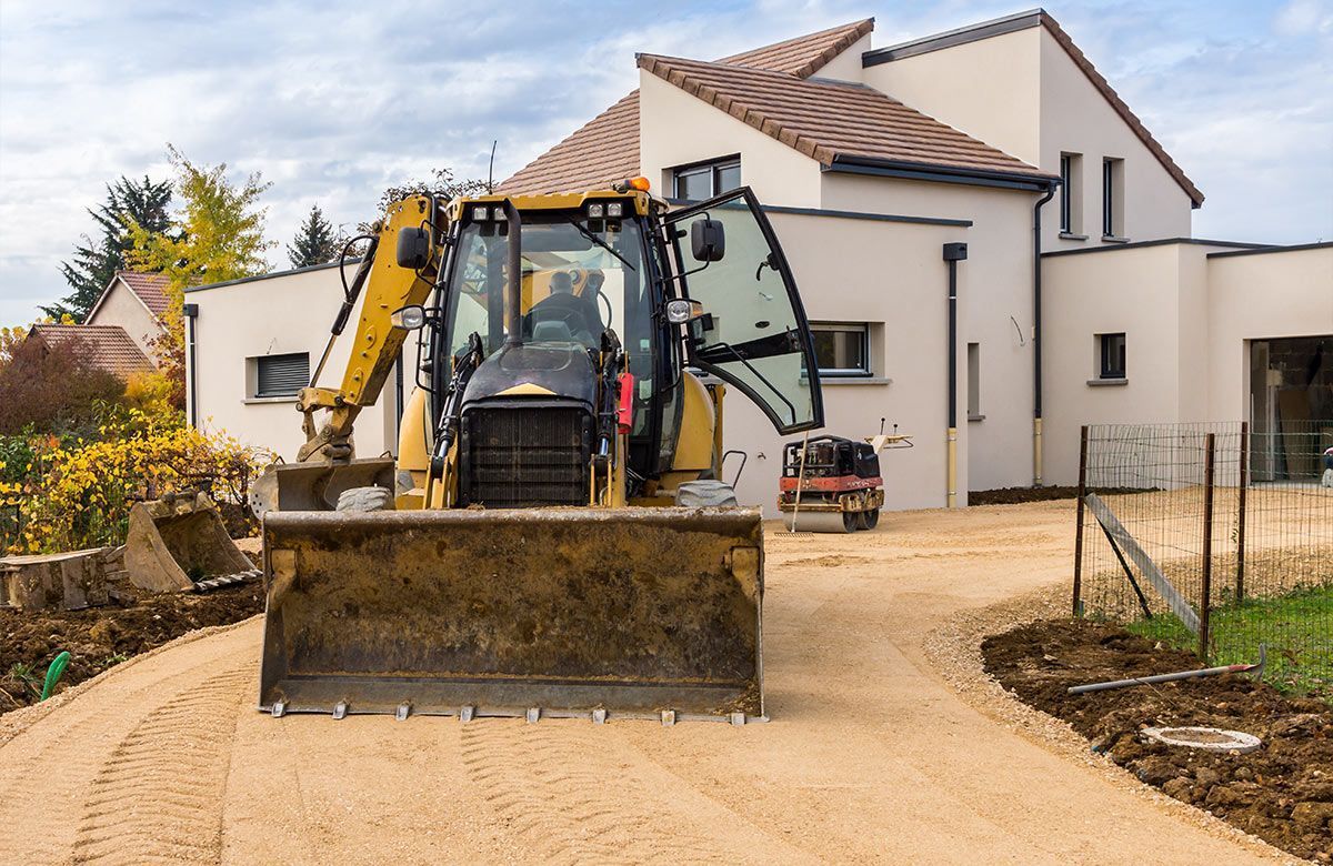 Une excavatrice nivelle une allée de gravier devant une maison moderne.