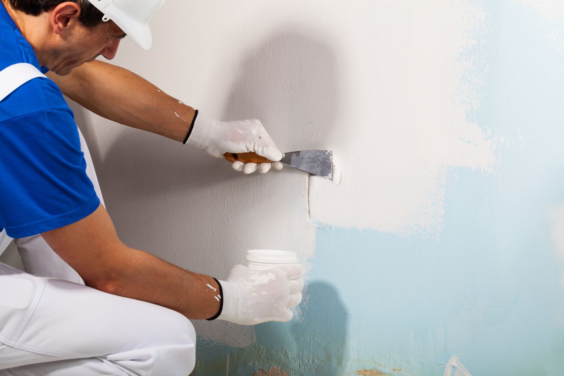 Ouvrier en chemise bleue et gants blancs appliquant un enduit sur un mur à la spatule.