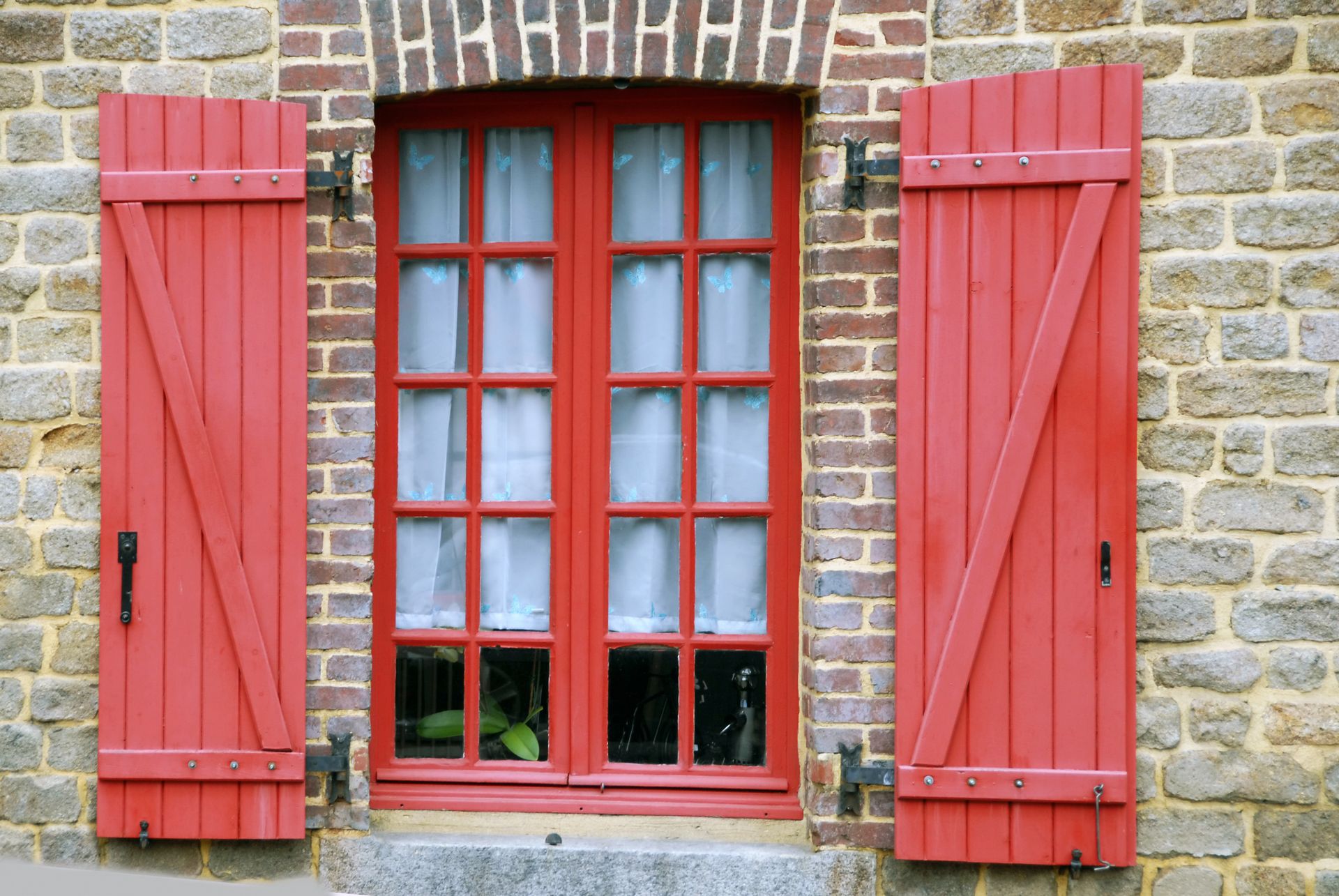 Fenêtre à volets rouges encastrée dans un mur de pierre, avec un cadre rouge et des rideaux blancs.