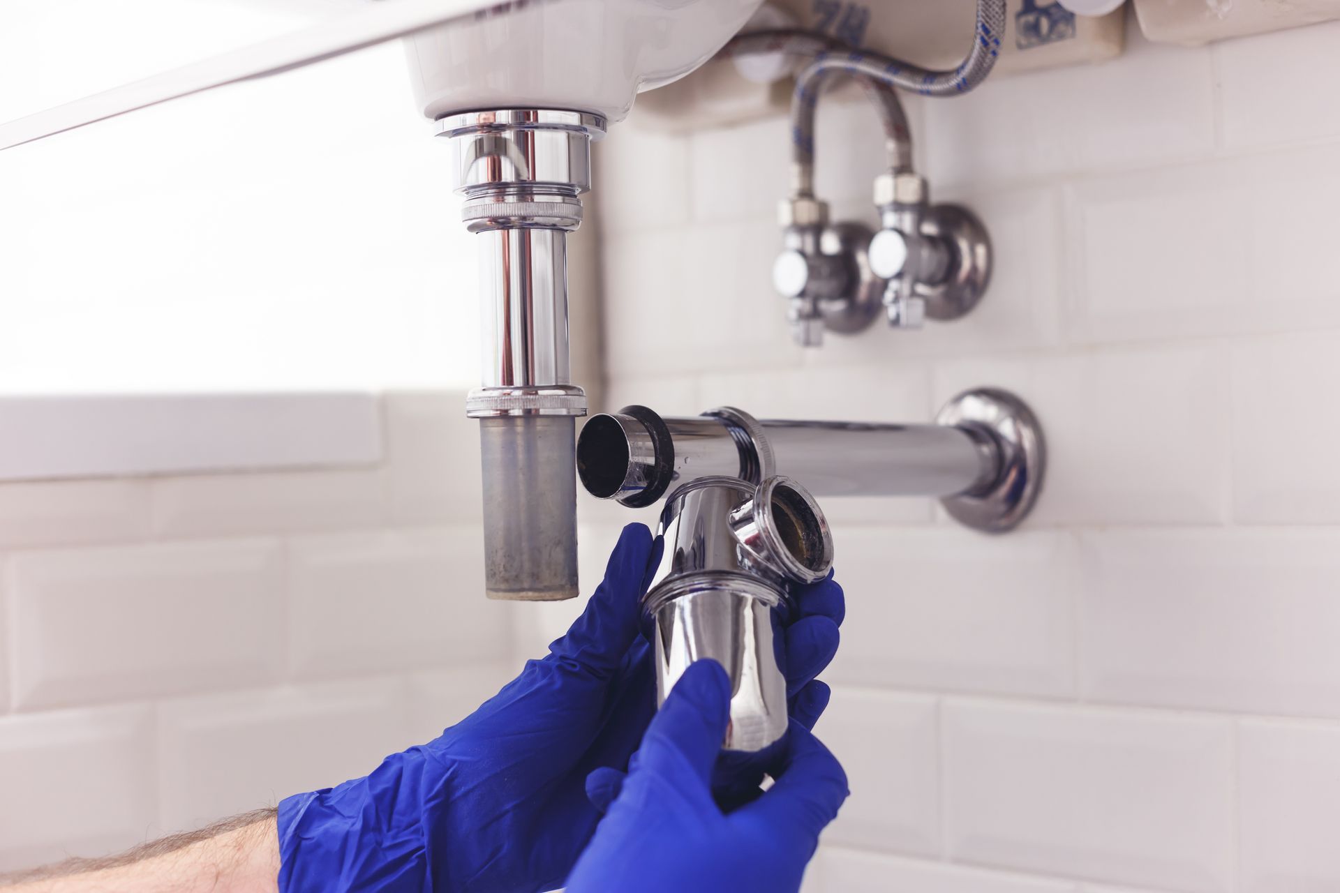Des mains gantées de bleu resserrent une bonde d'évier chromée dans une salle de bains carrelée de blanc.