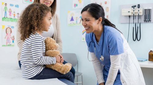 Un médecin sourit et discute avec un enfant tenant un ours en peluche dans une salle d'examen.
