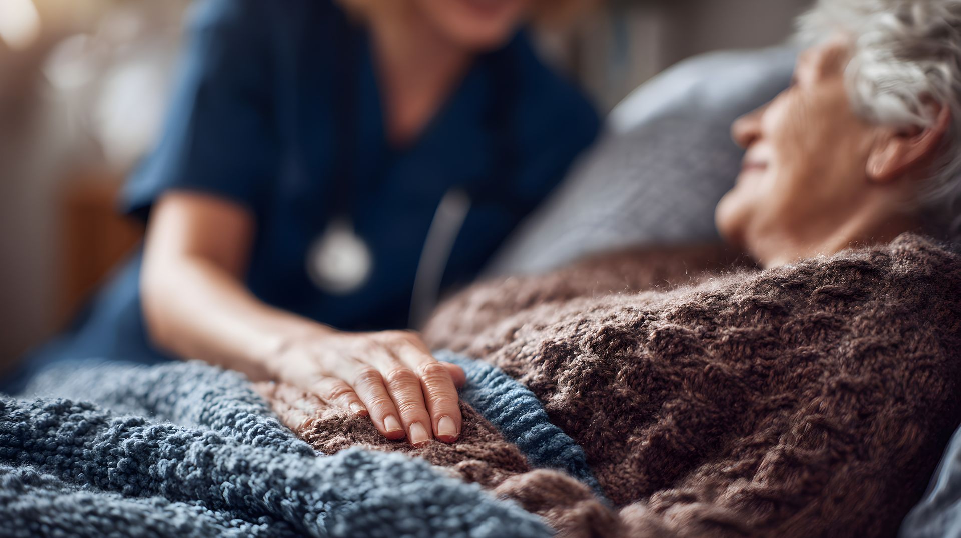 Une aide-soignante tient doucement la main d'un patient âgé allongé dans son lit sous une couverture bleue.