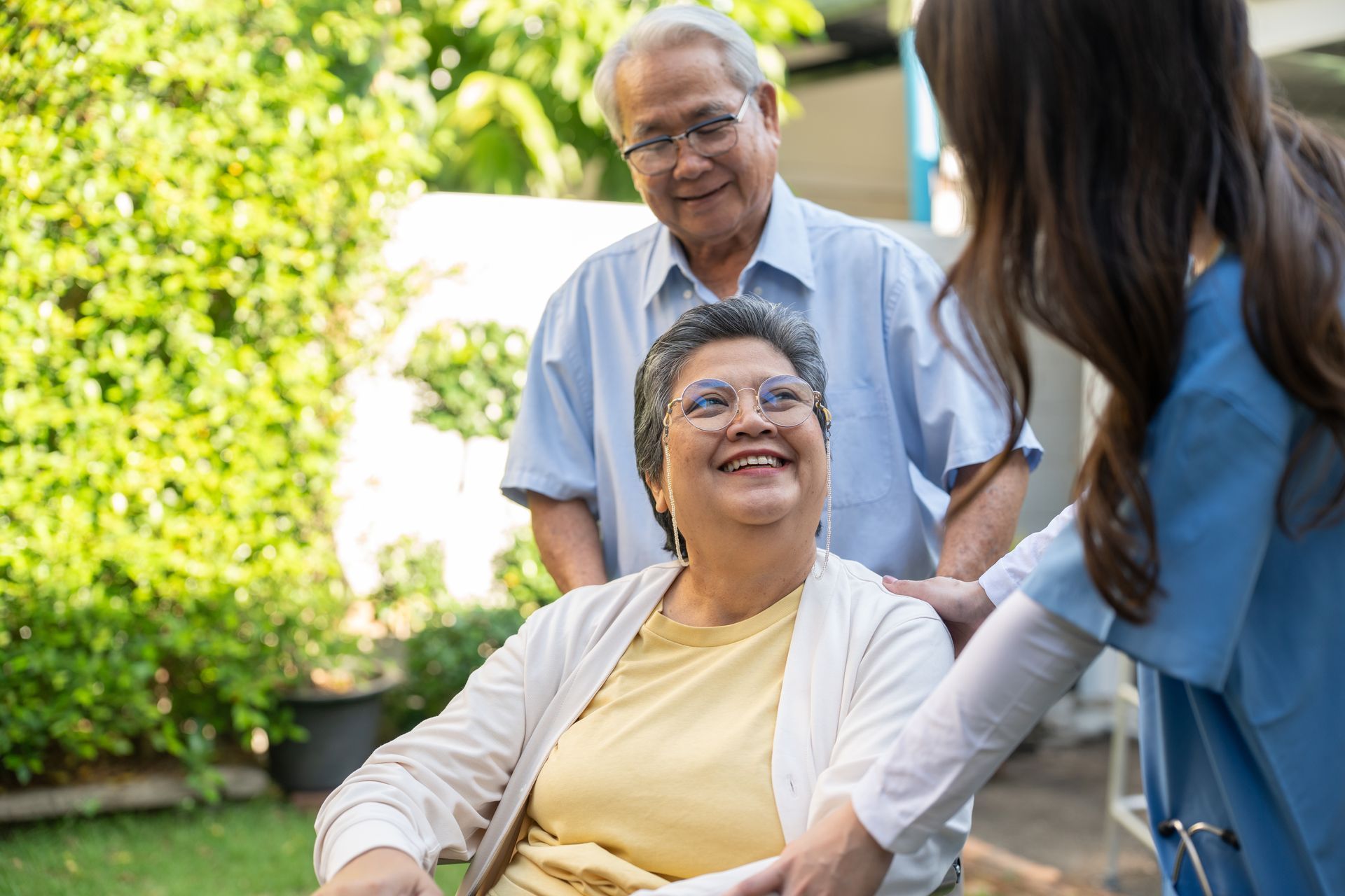 Une femme souriante en fauteuil roulant à l'extérieur, accompagnée d'un homme et d'une aide-soignante, dans un jardin.