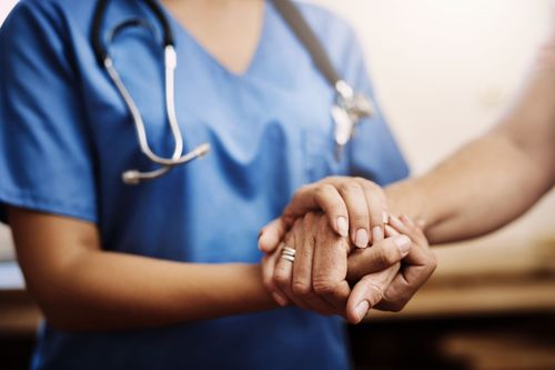 Une infirmière en blouse bleue tient délicatement la main d'un patient dans un environnement hospitalier