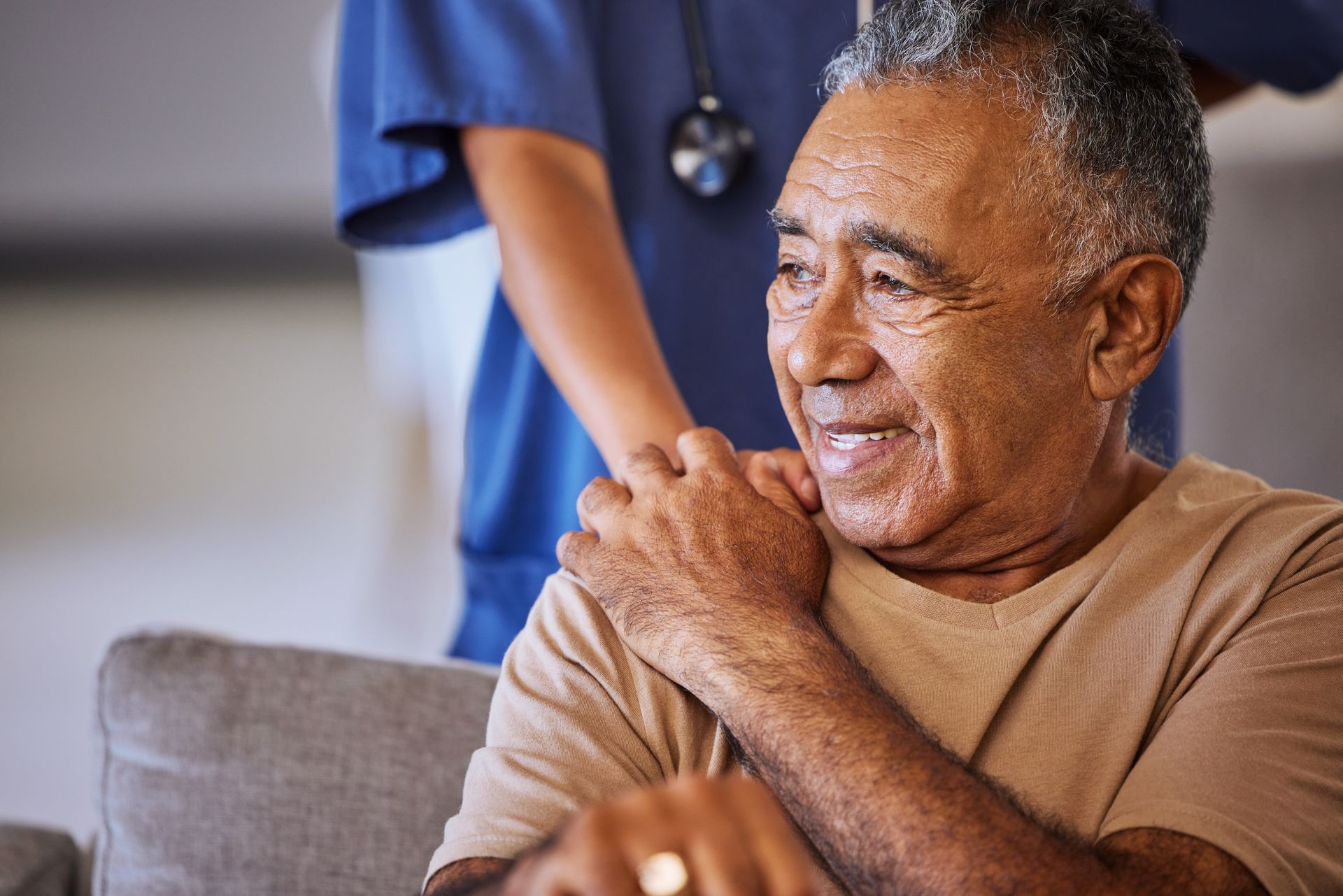 Un patient souriant est assis sur un canapé tandis qu'une infirmière se tient derrière lui, la main sur son épaule.