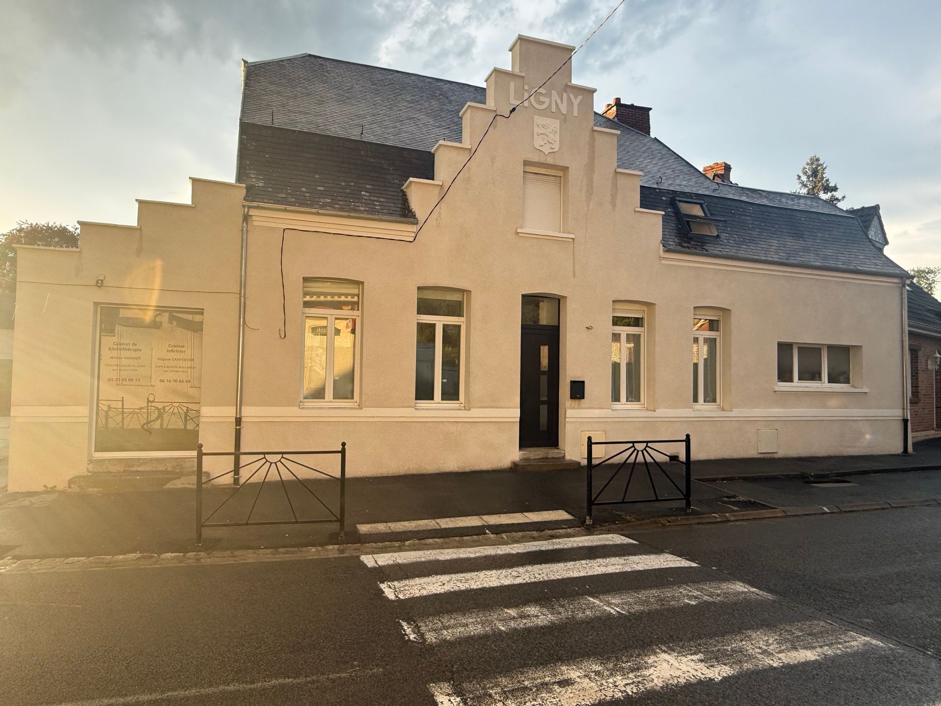 Bâtiment blanc au toit sombre et passage piéton rayé devant, éclairé par le soleil bas