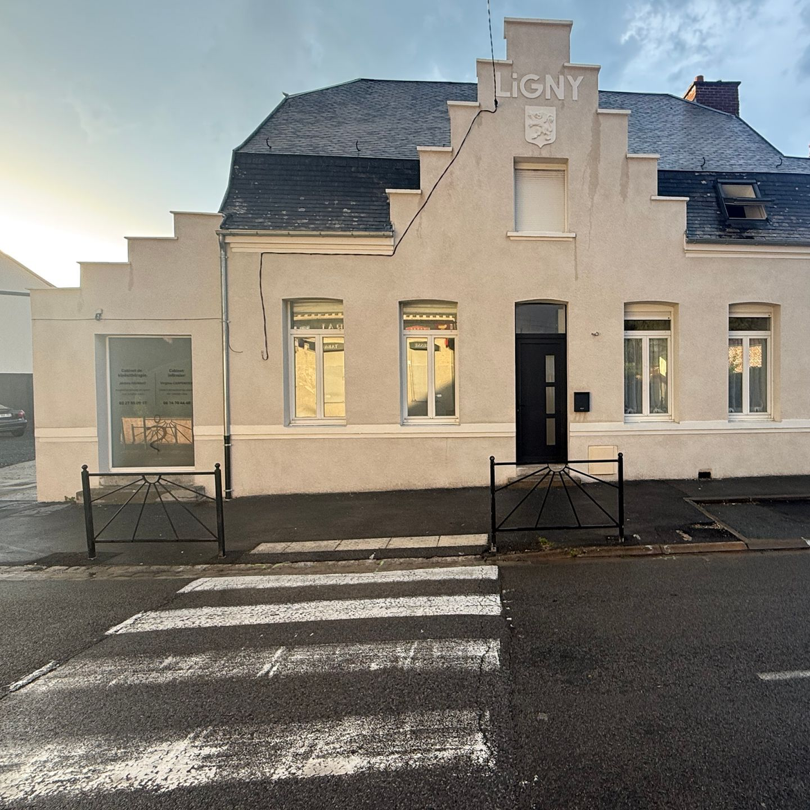 Bâtiment blanc avec entrée centrale et fenêtres, vu de l'autre côté d'une rue mouillée avec un passage piéton.