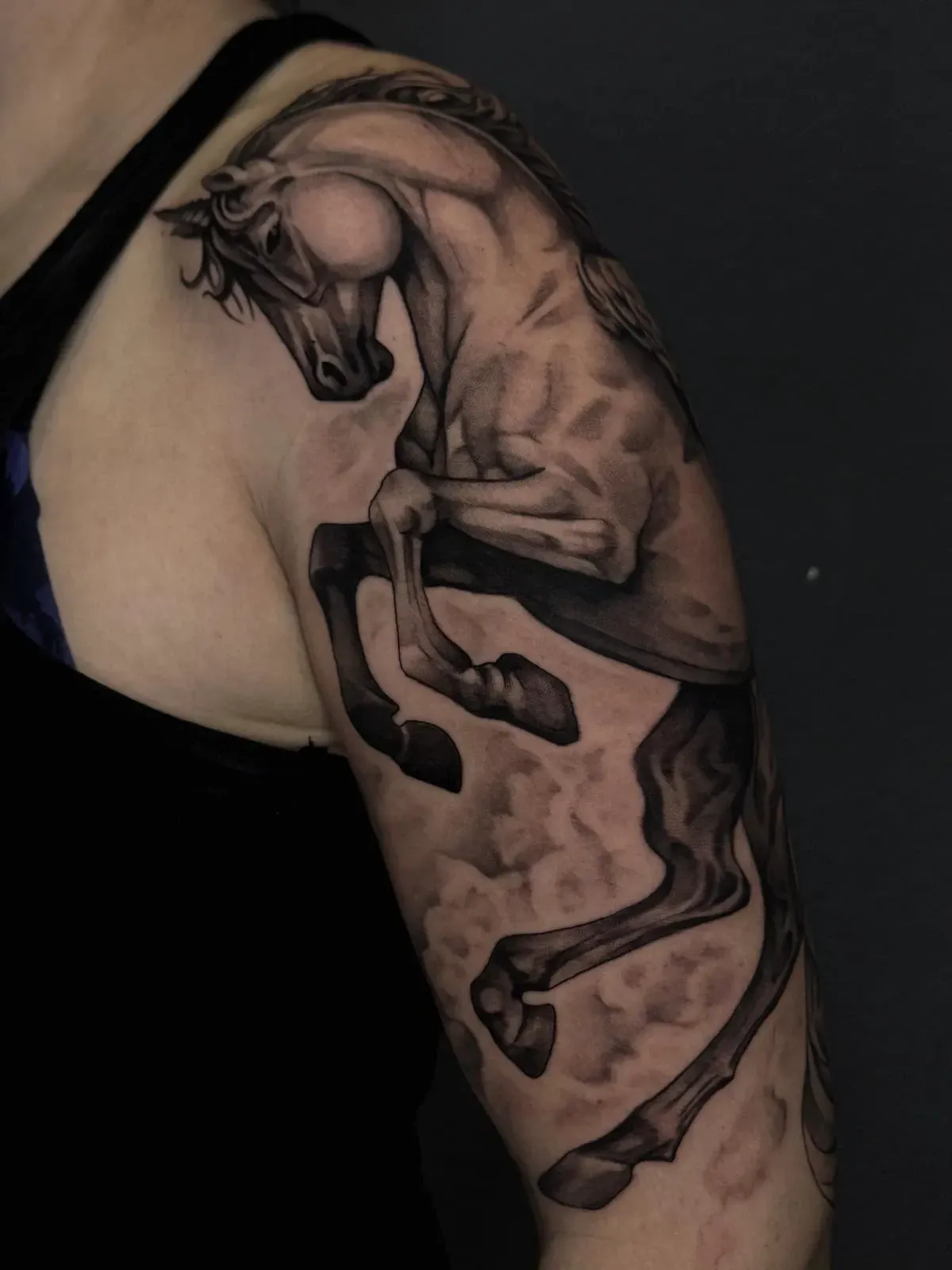 Tatuaje de un caballo negro y gris en la parte superior del brazo, encabritado y con sombras fluidas como el humo