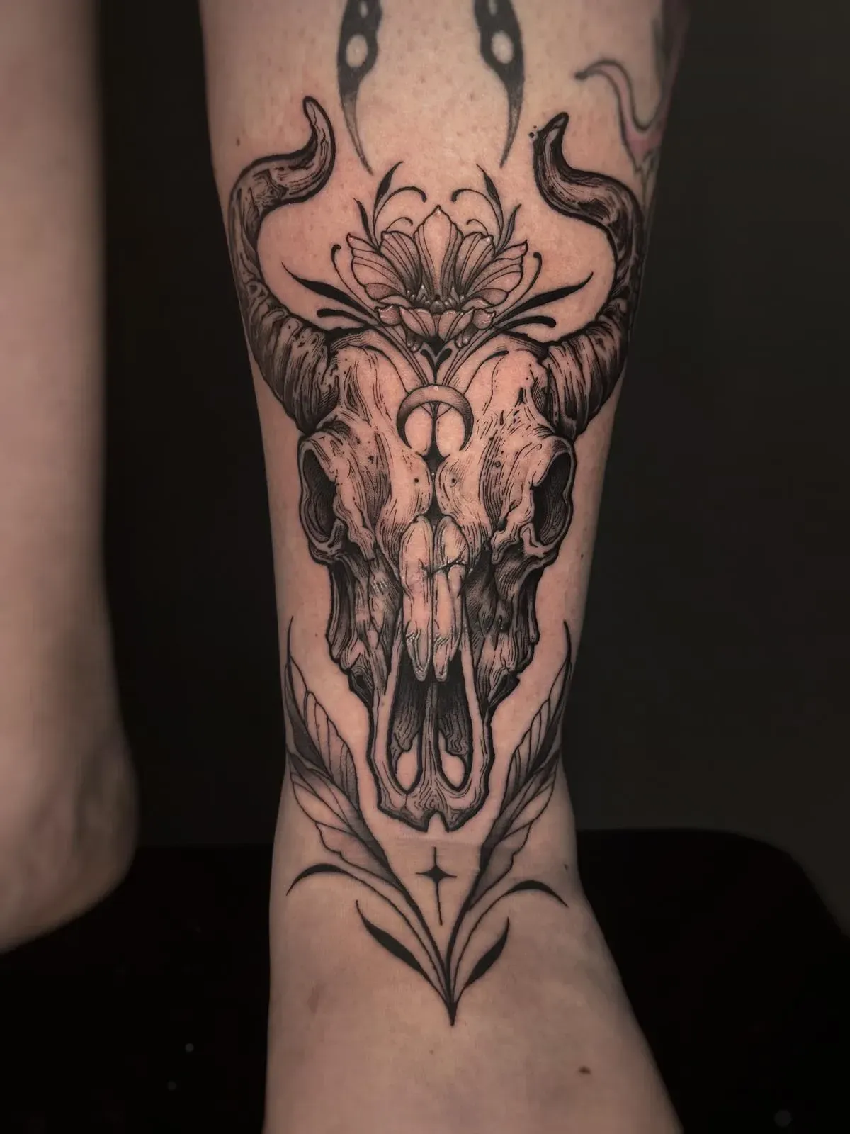 Tatuaje de una calavera en tinta negra en la pantorrilla, enmarcada por diseños ornamentados de flores