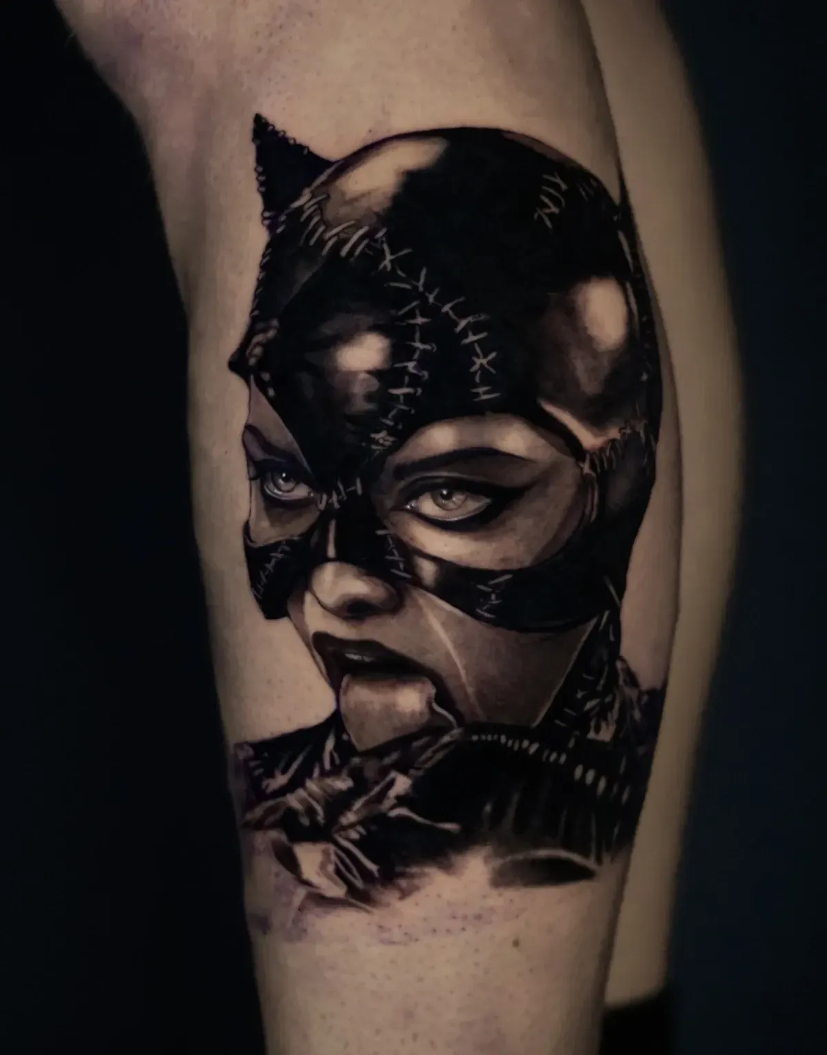 Tatuaje de Catwoman en negro y gris en un muslo, con una máscara llamativa, ojos y detalles de costuras