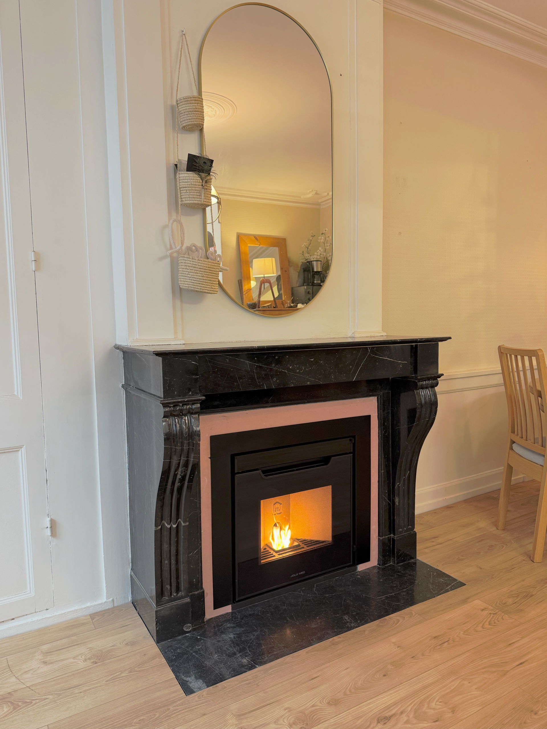 Cheminée noire avec un feu allumé, miroir ovale au-dessus, petites décorations.