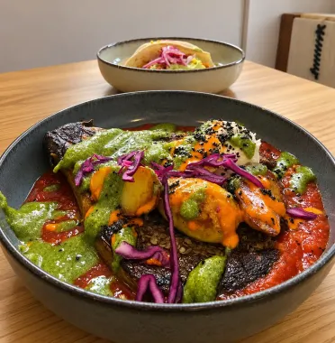 Plato gourmet colorido con salsa verde, verduras naranjas y col morada en un tazón negro sobre una mesa de madera.
